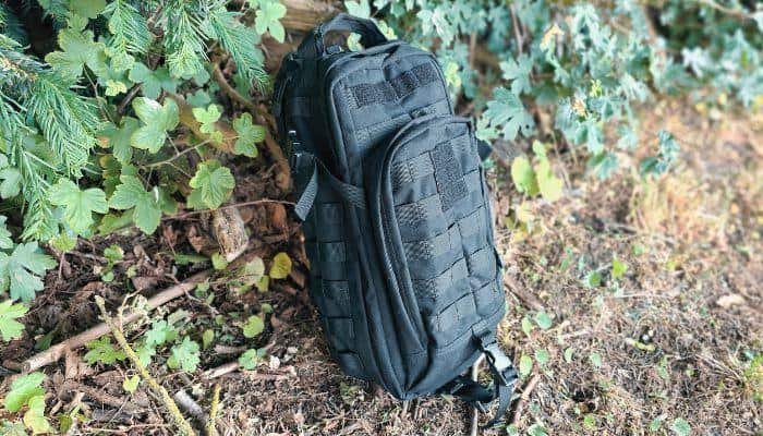 Backpack is laying on the ground, next to a tree. The brand of the bug out bag is the 5.11 Rush Moab 10